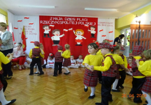 Dziewczynki w żółtych podkoszulkach i czerwonych spódniczkach w kratkę oraz chłopcy w żółtych koszulach, czarnych spodniach i czerwonych kamizelkach w kratkę tańczą w parach Polkę Bałucką.
